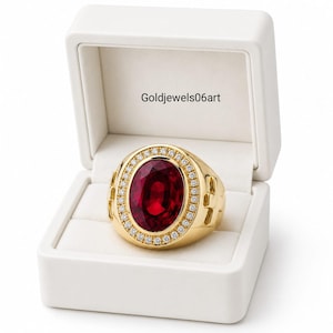 May include: A gold ring featuring a large oval-cut deep red gemstone surrounded by a halo of small clear stones, presented in a white box with the text Goldjewels06art printed on the interior lid. The ring has a polished yellow gold band.