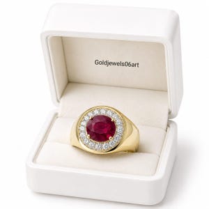 May include: A gold ring featuring a large, round, deep red ruby center stone surrounded by a halo of small, clear, sparkling diamonds. The ring is presented inside a white jewelry box with the text Goldjewels06art printed on the inner lid.