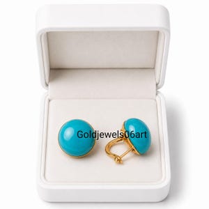 May include: A pair of round, smooth turquoise cabochon earrings set in gold-toned metal, featuring a hinged back closure. The earrings are displayed inside an open white jewelry box. The text Goldjewels06art is visible across the center.