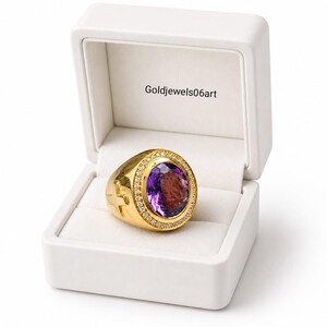 May include: A gold ring featuring a large oval-cut purple amethyst stone surrounded by a halo of small white stones. The band has a cross symbol on the side. The ring is presented in a white box with the text Goldjewels06art printed inside.