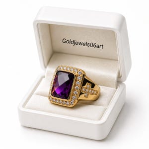 Puede incluir: Un anillo de oro con una gran piedra preciosa rectangular de color púrpura rodeada de pequeñas piedras claras, con un diseño de cruz en el lateral, presentado en una caja blanca con el texto Goldjewels06art impreso en la tapa interior.