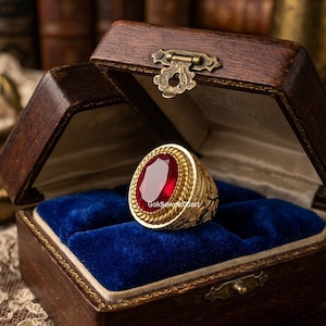 Pode incluir: Um anel de ouro com uma grande pedra vermelha de corte oval, inserida numa borda de corda torcida e uma banda gravada. O anel é exibido dentro de uma caixa de madeira escura com interior de veludo azul. O texto Goldjewels06art é visível no anel.