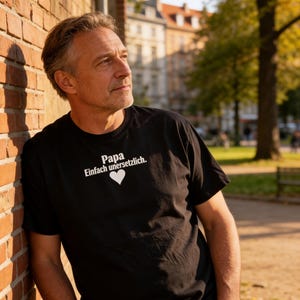 May include: A black t-shirt with the text "Papa Einfach unersetzlich." and a white heart graphic. The shirt is worn by a person leaning against a brick wall. The background includes trees and buildings.