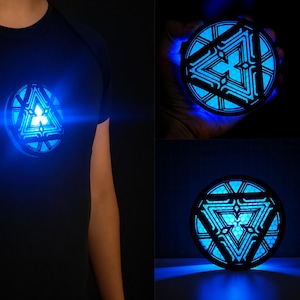 Könnte beinhalten: Ein kreisförmiges, schwarzes Requisit im mechanischen Stil mit einem leuchtend blauen, dreieckigen Lichtmuster und komplexen geometrischen Ausschnitten. Das Gerät strahlt helles, kühles blaues Licht aus und ist an einem dunklen Shirt befestigt.