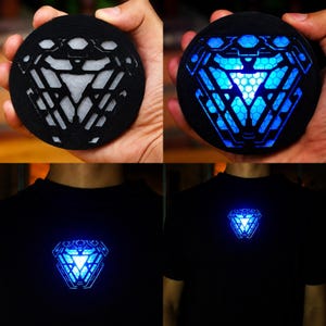 Può includere: Un reattore ad arco circolare nero con un design geometrico ritagliato e uno strato interno bianco. Il dispositivo è dotato di una luce LED blu brillante che illumina il motivo esagonale dall'interno. È mostrato sia spento che acceso su una maglietta nera.