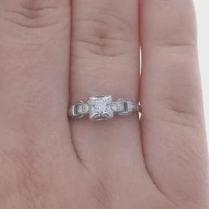 May include: A silver-toned metal ring featuring a central square-shaped setting with a small embedded stone. The band has a geometric design with rectangular accents on either side of the center, creating a structured and symmetrical appearance.