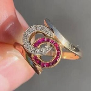 Puede incluir: Un anillo de oro con dos motivos circulares entrelazados. Un círculo está engastado con pequeñas piedras claras, mientras que el otro círculo está engastado con piedras rojas vibrantes. La banda del anillo es lisa y pulida, unida a los elementos.