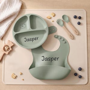 Può includere: Un set pappa per bambini verde salvia con il nome "Jasper" stampato sul piatto e sul bavaglino. Il set include un piatto diviso, un bavaglino, una forchetta e due cucchiai con manici in legno. Sono presenti anche un sonaglio di legno e mirtilli.