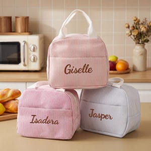 May include: Three personalized lunch bags in pink, red, and gray with white handles. The bags have vertical stripes and the names "Giselle", "Isadora", and "Jasper" embroidered on them. The bags are sitting on a kitchen counter.