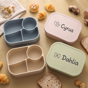 Puede incluir: Cuatro fiambreras de colores con compartimentos y tapas. Dos cajas son de color azul claro y beige, y dos son rosas y verdes. La caja rosa tiene impreso "Cyrus", y la caja verde tiene impreso "Dahlia".