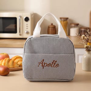 May include: A black and white striped lunch bag with a white handle and the embroidered word "Apollo" in brown. The bag has a front pocket and is sitting on a wooden surface. A microwave and food items are in the background.