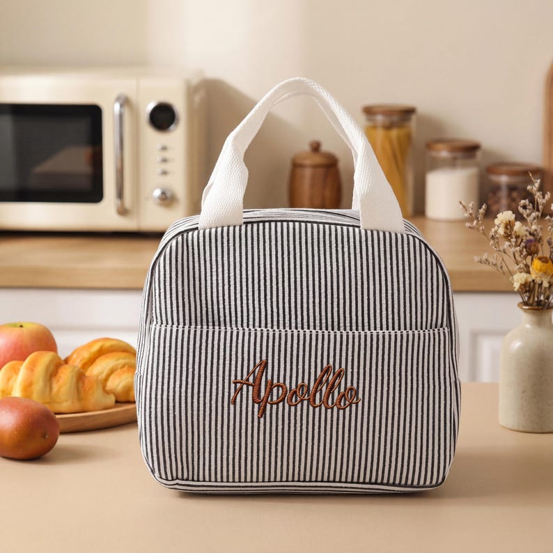 May include: A black and white striped lunch bag with a white handle and the embroidered word "Apollo" in brown. The bag has a front pocket and is sitting on a wooden surface. A microwave and food items are in the background.