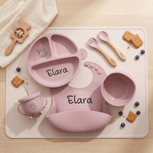 May include: A baby feeding set in a soft pink color, including a divided plate, bowl, bib, cup, spoon, and fork. The name "Elara" is printed on the plate and bib. A wooden toy and cookies are also present.