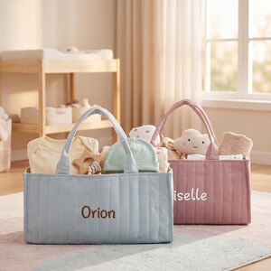 May include: A light blue quilted fabric storage basket with the name Orion embroidered in brown, and a dusty pink quilted fabric storage basket with the name Giselle embroidered in white, both filled with baby clothing and accessories.