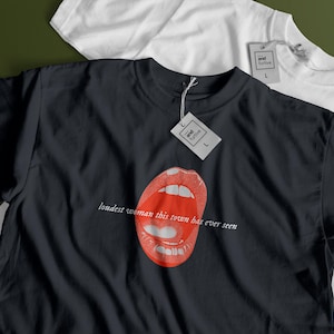 May include: Black t-shirt with a graphic of red lips and the text "loudest woman this town has ever seen." A white t-shirt is visible in the background. Both shirts have tags attached.