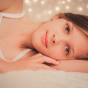 May include: A young person with long brown hair lies on a white surface with their head resting on their arm. They are wearing a white vest top and have their eyes open, looking directly at the camera.