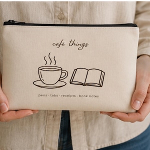 以下が含まれることがあります： 黒いジッパーが付いたベージュ色のキャンバス地のポーチ。 「café things」の文字と、湯気の立つコーヒーカップと開いた本のイラストが描かれています。 イラストの下には「pens · tabs · receipts · book notes」の文字があります。