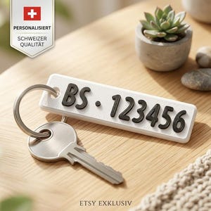 Op de afbeelding: Een rechthoekige witte sleutelhanger met verhoogde zwarte letters die BS 123456 vormen, bevestigd aan een zilveren metalen ring met een zilveren sleutel. Het label heeft een strak ontwerp met een gladde afwerking en afgeronde hoeken.