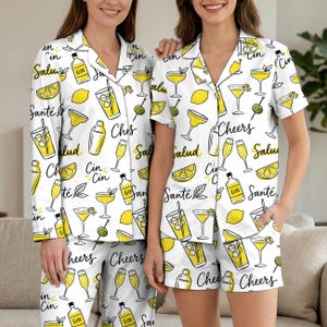 Könnte beinhalten: Weißes Pyjama-Set mit einem sich wiederholenden Muster aus gelben Cocktail-Illustrationen, Zitronen und Text. Das Set beinhaltet ein langärmliges Hemd mit Knöpfen und eine Hose sowie ein kurzärmliges Hemd und Shorts.