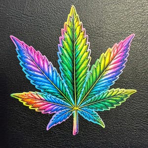 Puede incluir: Una colorida pegatina de una hoja de cannabis. La hoja es una vibrante mezcla de colores del arcoíris, incluyendo azul, rosa, amarillo y verde. La pegatina está sobre un fondo negro texturizado. Las venas de la hoja están detalladas con líneas amarillas.