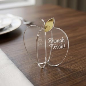 May include: A clear acrylic apple-shaped ornament featuring a gold-coloured leaf accent at the top. The surface of the acrylic is engraved with the white text "Shanah Tovah!" in a script font, creating a three-dimensional structure with intersecting panels.