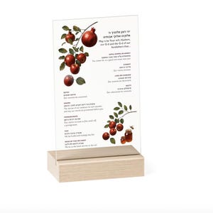 May include: A clear acrylic sign with a light wood base featuring illustrations of pomegranates and honey, along with printed text in English and Hebrew detailing traditional Rosh Hashanah blessings for foods like apples, carrots, and fish.