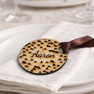 May include: A wooden place card with the name "Aaron" laser-cut into it, featuring a unique dotted pattern. The place card is attached to a brown ribbon and rests on a white napkin, placed on a white plate. The setting suggests a formal dining occasion.