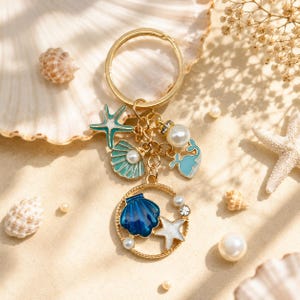 May include: Gold-tone keychain with ocean-themed charms. The charms include a blue seashell, starfish, and pearls. The keychain is adorned with a round charm featuring a blue seashell and a white starfish. The background includes seashells and pearls.