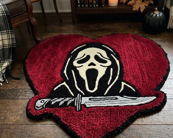 May include: A heart-shaped tufted rug featuring a deep red background with a black and white graphic of a masked figure holding a silver knife. The rug has a thick, textured pile and a black border, showcasing a detailed horror-themed design.