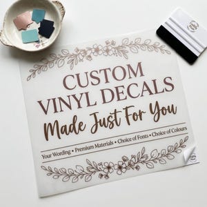 Może przedstawiać: Biała kartka z brązowym napisem "CUSTOM VINYL DECALS Made Just For You". Arkusz ma kwiatowe wzory i tekst "Your Wording • Premium Materials • Choice of Fonts • Choice of Colours". Obecna jest również biała rakla i miska z próbkami kolorów.