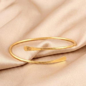 May include: A gold-toned metal bangle bracelet featuring a bypass design with two slightly flattened, rounded ends. The bracelet has a smooth, polished finish and a circular shape that remains open, allowing for adjustment around the wrist.