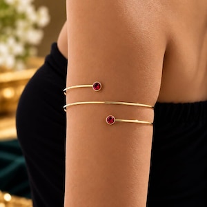 May include: A gold-toned metal arm cuff featuring a spiral design that wraps around the upper arm. The two ends of the wire are each finished with a small, round, vibrant red gemstone set in a simple gold-toned bezel setting.