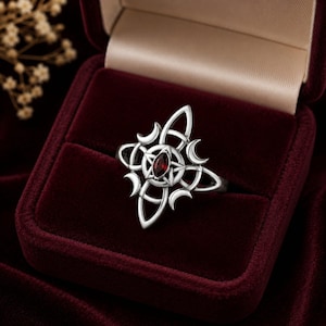 Peut inclure: Une bague en argent avec une pierre précieuse rouge foncé de taille marquise sertie au centre d'une étoile à cinq branches. L'étoile est entourée d'une structure métallique complexe de boucles entrelacées et de quatre croissants de lune.