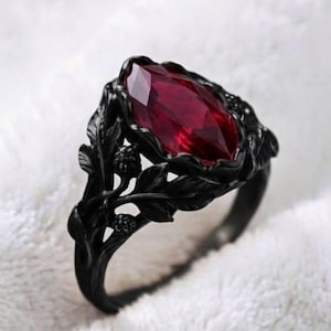 May include: A black metal ring featuring a marquise-cut deep red gemstone at the centre. The band is designed with intricate, sculpted black leaves and small berry-like textures that wrap around the finger, framing the central red stone.