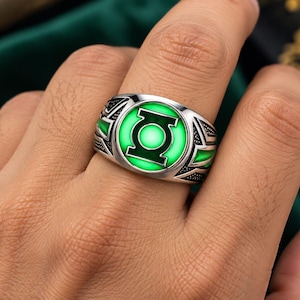 May include: A silver-toned ring featuring a circular centrepiece with a bright green glowing emblem of a stylised lantern symbol. The band has intricate, dark-etched geometric patterns on the sides, providing a textured contrast to the smooth, polished metal finish of the ring's main structure.