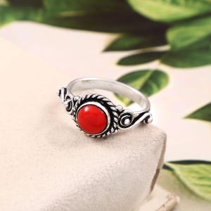 Könnte beinhalten: Ein silberner Ring mit einem zentralen, runden, glatten und leuchtend roten Stein. Das Metallband weist eine gedrehte Seilstruktur um den Stein herum auf, mit dekorativen Schnörkeldetails, die sich entlang der Seiten des Bandes erstrecken.