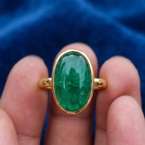 May include: A gold ring featuring a large, oval-shaped, cabochon-cut green gemstone set in a smooth, polished gold bezel. The ring band is thin and circular, crafted from the same yellow-toned metal as the setting, creating a simple, elegant design.