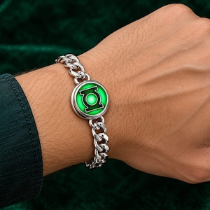 Könnte beinhalten: Ein silberfarbenes Gliederkettenarmband aus Metall mit einem runden Anhänger, der in der Mitte ein leuchtendes grünes und schwarzes Green Lantern-Logo zeigt. Das Armband besteht aus polierten, ineinandergreifenden Gliedern und einer runden Fassung.