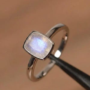 May include: A silver-toned ring featuring a square-shaped moonstone with rounded corners set in a smooth bezel setting. The gemstone displays a translucent appearance with a soft blue iridescent glow, mounted on a polished, plain metal band.