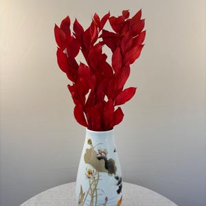 Pode incluir: Um vaso de cerâmica branca com pescoço afunilado e corpo arredondado, decorado com ilustrações pintadas de flores de lótus, vitórias-régias e peixes dourados. O vaso está preenchido com um ramo de folhas de ruscus preservadas em vermelho vivo.