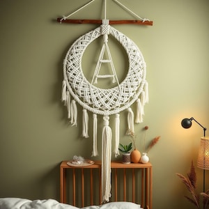 Può includere: Un grande arazzo da parete in macramè bianco sporco con un design circolare e una struttura a forma di scala al centro. Il macramè è sospeso da un tassello di legno e presenta più nappe. L'arazzo è esposto su una parete verde tenue.