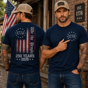 Op de afbeelding: Een marineblauw t-shirt met een verweerde Amerikaanse vlag op de achterkant met de tekst We the People, 1776 en 250 YEARS 2026. De voorkant van het shirt toont een kleine cirkelvormige afbeelding met 1776 omringd door dertien sterren.