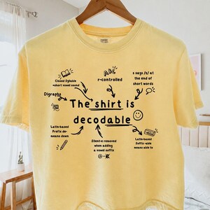 Op de afbeelding: Een lichtgeel t-shirt met korte mouwen en zwarte tekst en illustraties. De centrale tekst luidt, The shirt is decodable, omringd door educatieve taalkundige notities zoals r-controlled, digraphs en silent-e, met een Comfort Colors label.