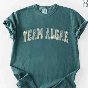 Könnte beinhalten: Ein kurzärmeliges T-Shirt in Blaufichtengrün mit Rundhalsausschnitt. Auf der Brust befindet sich der Schriftzug "TEAM ALGAE" in einer abgenutzten, cremeweißen College-Schriftart. Das Kleidungsstück hat ein Comfort Colors Markenetikett am Innenkragen.