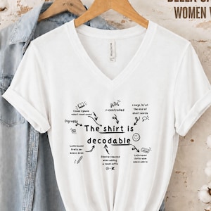 Op de afbeelding: Een wit T-shirt met V-hals voorzien van zwarte tekst en grafische illustraties. De centrale tekst luidt The shirt is decodable, omringd door kleine diagrammen en labels waaronder r-controlled, digraphs en Latin-based prefix means down.