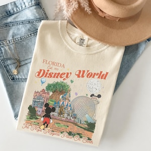 May include: Cream-colored t-shirt with a Disney World graphic, jeans, a gold necklace, and a tan hat. The shirt features the words "Florida Disney World" and a colorful illustration of the theme park. The jeans are light blue.