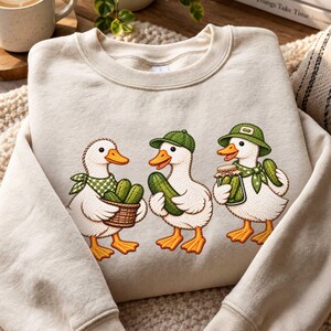 Peut inclure: Un sweat-shirt à col rond couleur crème orné d'une broderie représentant trois canards blancs. Les canards portent des accessoires verts et tiennent des cornichons, l'un portant un panier de cornichons et un autre tenant un bocal en verre.