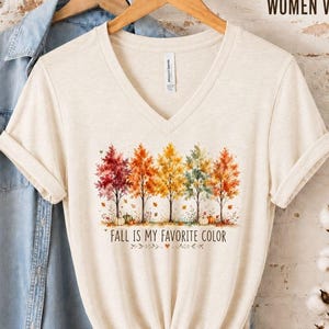May include: A cream-coloured V-neck t-shirt featuring a graphic of five autumn trees in shades of red, orange, and yellow with the text FALL IS MY FAVOURITE COLOR. The shirt is displayed on a wooden hanger alongside a light blue denim shirt.