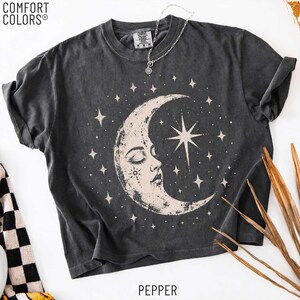 May include: A charcoal gray short-sleeve t-shirt with a crew neckline and a graphic print of a crescent moon with a human face surrounded by stars. The shirt features the Comfort Colors brand label at the neck and the text Pepper at the bottom.