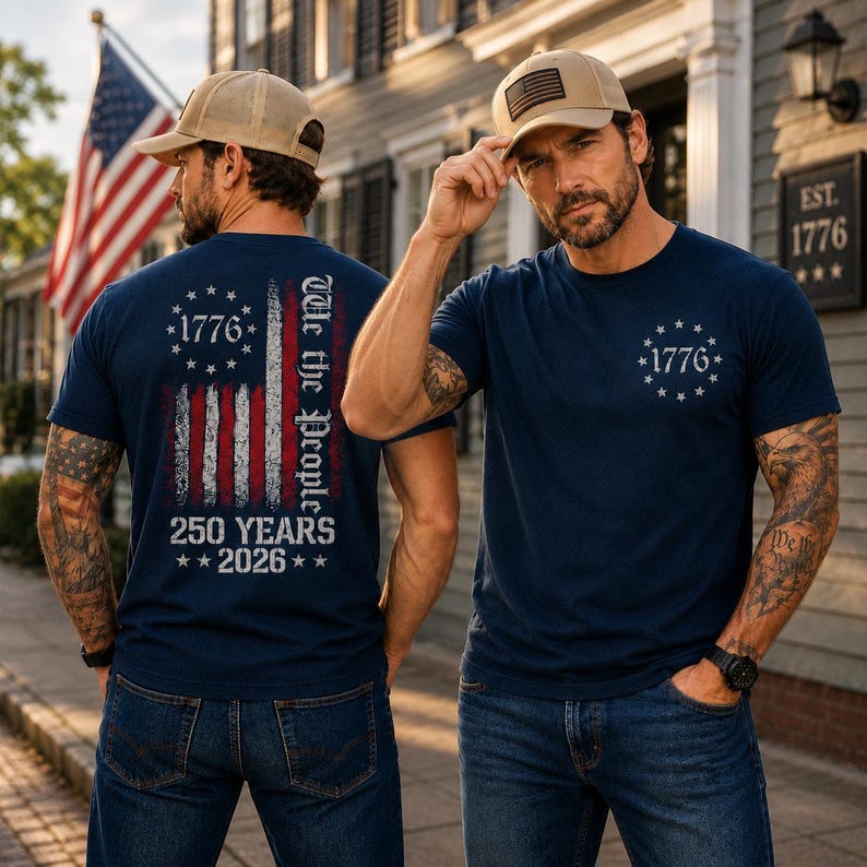 Op de afbeelding: Een marineblauw t-shirt met een verweerde Amerikaanse vlag-afbeelding, de tekst "We the People", "1776" en "250 YEARS 2026" op de achterkant, en een "1776" ster-embleem op de borst, gecombineerd met een beige baseballpet met een vlag-patch.